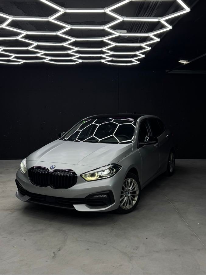 BMW Serie 1 con Car Wrap gris satinado, tres cuartos delantero bajo las luces hexagonales del estudio