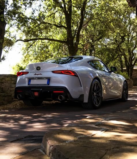White Toyota GR Supra rear three-quarter parked under trees on a stone driveway